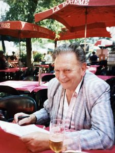 Jacques Devillers sonriente en una terraza de París con papeles de trabajo.